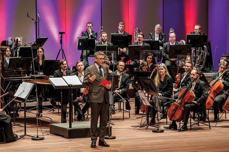 Een dirigent staat op het podium voor een orkest en spreekt het publiek toe, met muzikanten op de achtergrond.