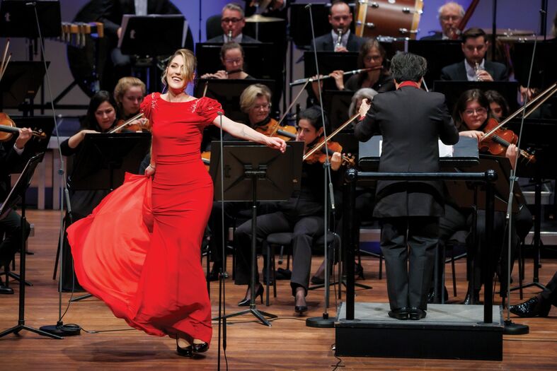 Vrouw in een rode jurk zingt en danst voor een orkest, terwijl de dirigent voor de muzikanten staat.