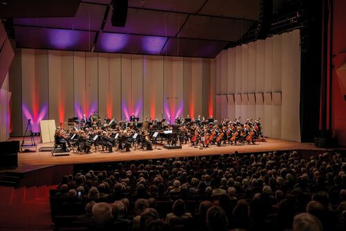 Een orkest speelt op het podium van een concertzaal, met een publiek in de voorgrond en kleurrijke verlichting.
