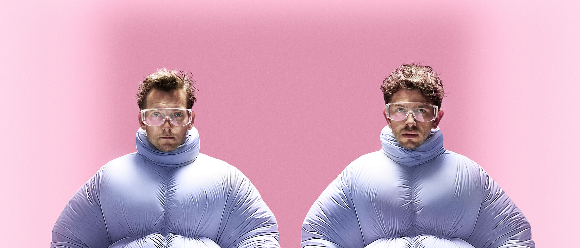 Twee personen in opgeblazen lichtblauwe pakken rennen synchroon naar voren tegen een roze achtergrond.