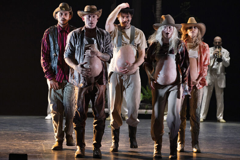 Vijf performers in cowboyoutfits met nep-buiken, staan op een podium en kijken in de richting van het publiek.