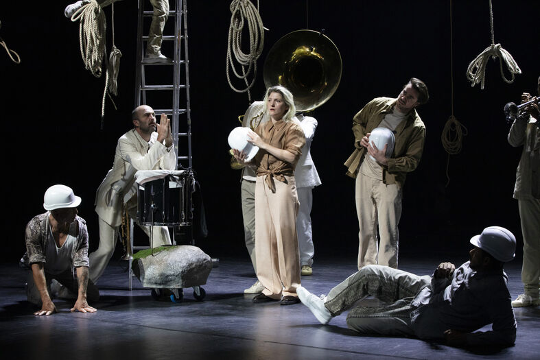 Theaterperformance met acteurs in verschillende poses, met rekwisieten zoals ladders en schalen, op een donkere achtergrond.