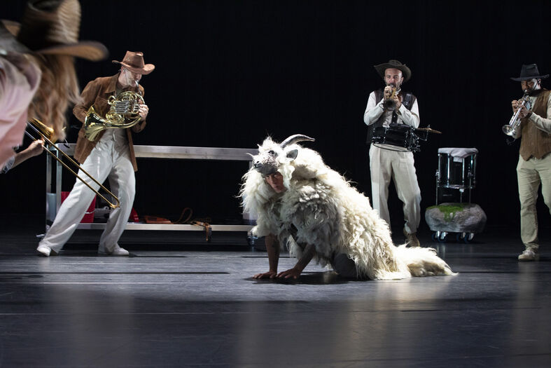 Performers op het podium met een grote, harige pop en muzikanten op de achtergrond, in een theatrale setting.
