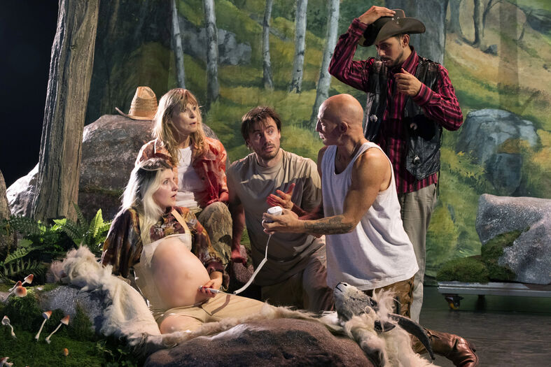 Een groep mensen in kostuums voert een theatrale scène op in een bosachtige setting, met een vrouw in het midden.