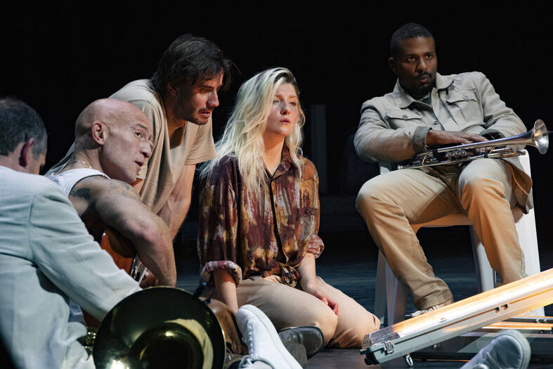 Theaterafbeelding van vijf acteurs in een dynamische scène, zittend en gefocust op elkaar.