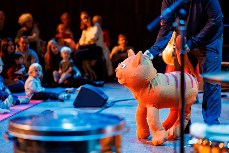 Een groot, vrolijk oranje knuffeldier in de vorm van een kat wordt gepresenteerd op een podium voor een publiek van kinderen.