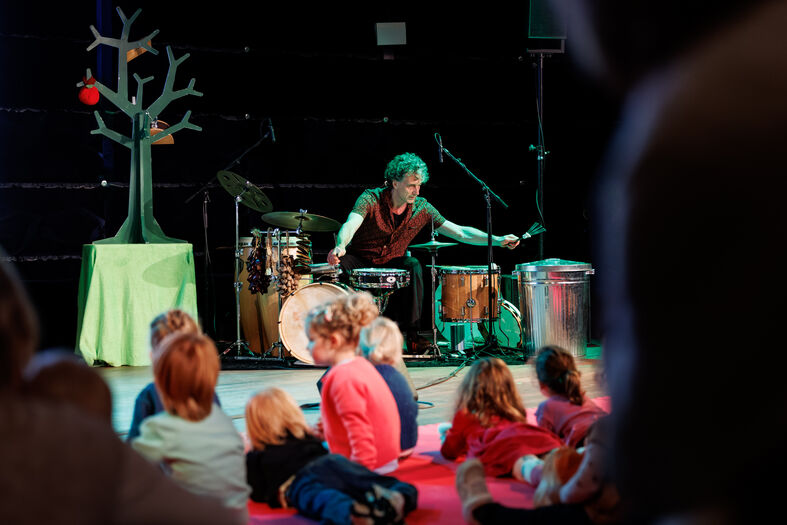 Een muzikant speelt op percussie-instrumenten voor een groep kinderen die op de vloer zitten.