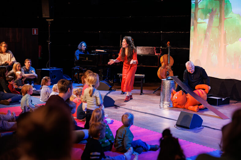 Een verteller en muzikanten voeren een optreden uit voor kinderen in een theateromgeving.