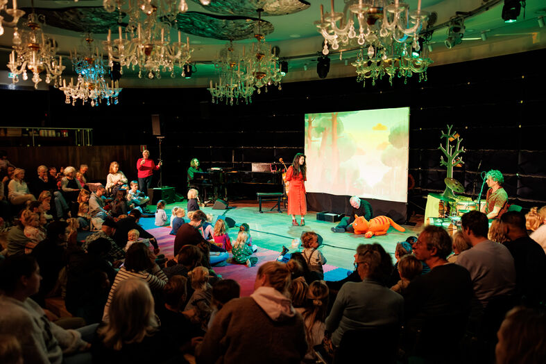 Een podium met performers en een projector, omringd door een publiek dat actief kijkt en geniet van de voorstelling.