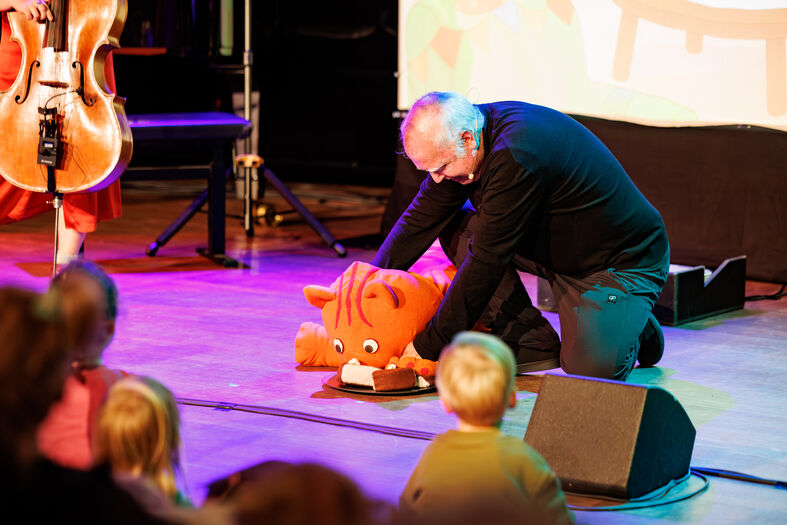 Man knielt op het podium naast een grote, pluchen oranje vis, terwijl kinderen in de zaal toekijken.