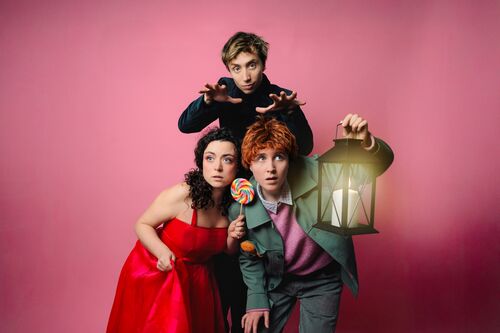 Drie mensen poseren op een roze achtergrond; één houdt een lantaarn vast, terwijl de anderen naar de camera kijken.