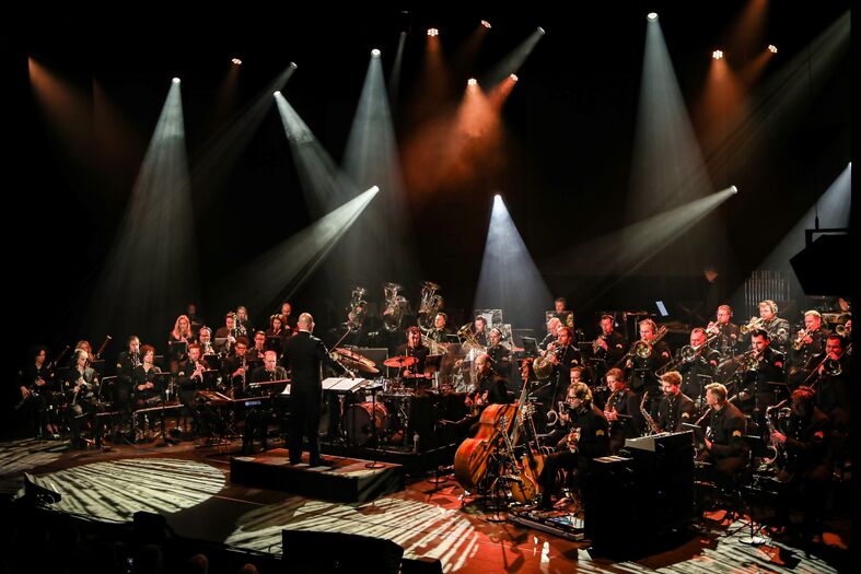 Een orkest speelt op een podium, verlicht door verschillende lichtstralen, met een dirigent vooraan.