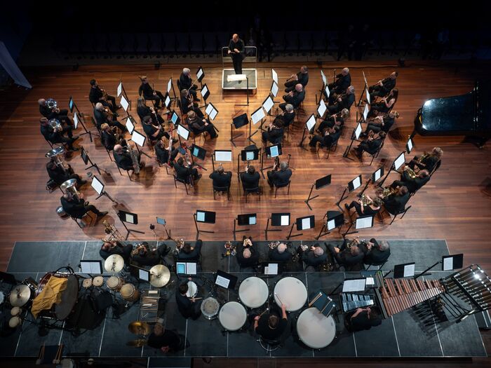 Bovenaanzicht van een orkest dat repeteert, met diverse instrumenten en muzikanten in een concertzaal.