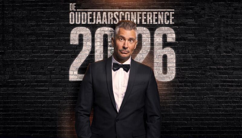 Guido Weijers in een smoking voor een donkere stenenmuur.