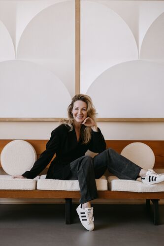 Debby Gerritsen zit ontspannen op een bank met lichte ronde kussens, terwijl een ander op de leuning leunt in een modern interieur met geometrisch reliëf aan de wand.