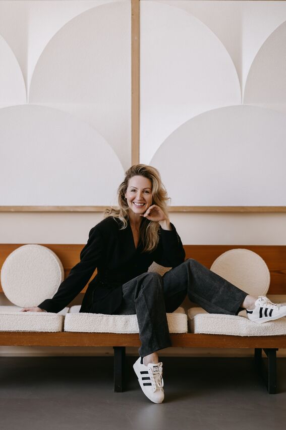 Debby Gerritsen zit ontspannen op een bank met lichte ronde kussens, terwijl een ander op de leuning leunt in een modern interieur met geometrisch reliëf aan de wand.