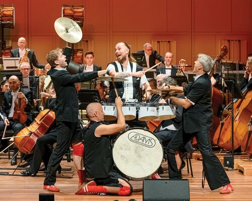 Drie muzikanten voeren een energieke act uit op het podium, met een orkest op de achtergrond.