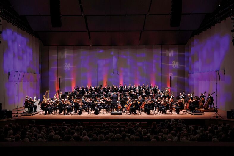 Groot orkest en koor treden op onder gekleurde podiumverlichting voor publiek.