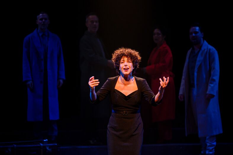 Zangeres op het podium met dramatische verlichting, omringd door schimmige figuren in achtergrond.