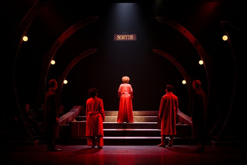 Een vrouw in een rode jurk staat op een trap, omringd door figuren in schaduw, met een spotlicht boven haar.