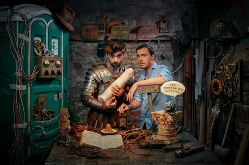 Twee mannen in een middeleeuwse werkplaats, één in harnassing met een rol perkament, de ander verdiept in een boek.