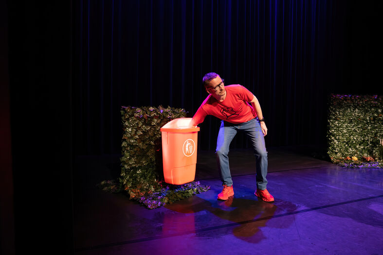 Een man in een rode shirt gooit iets in een rode prullenbak op een podium, omringd door groene hagen.