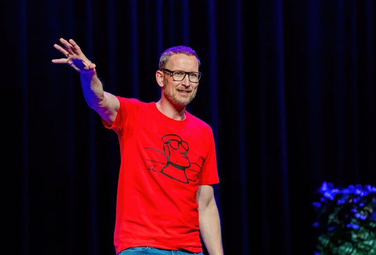 Man in een rode T-shirt spreekt op een podium met een donkere achtergrond en groene planten.