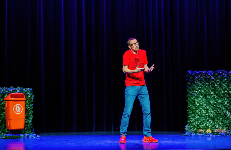Man in rode T-shirt en blauwe jeans staat op een podium, met een vuilnisbak en groene planten op de achtergrond.