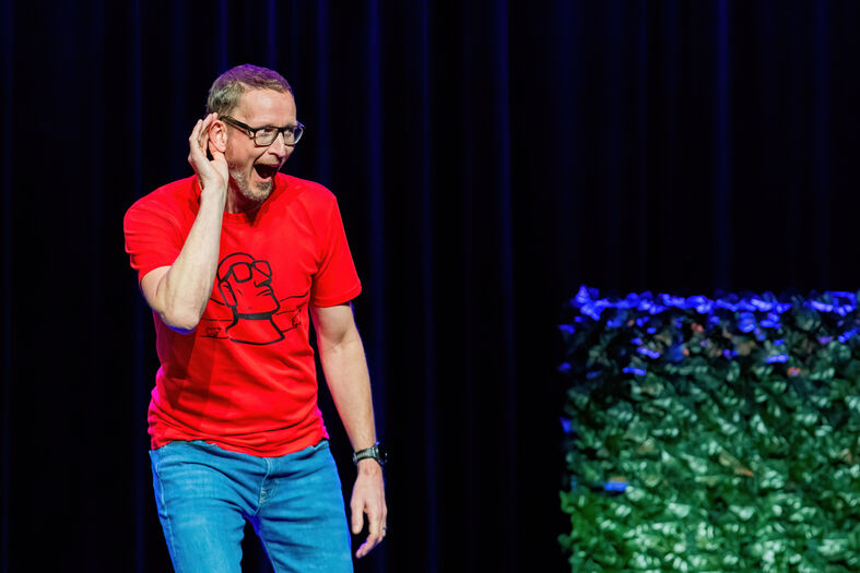 Een man in een rode shirt staat op een podium en houdt zijn hand aan zijn oor, lijkt te luisteren.