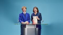 Twee vrouwen in formele kleding staan achter een glazen tafel op een blauwe achtergrond, lachend en met documenten in hun handen.