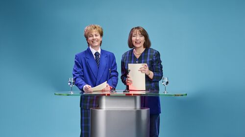 Twee vrouwen in formele kleding staan achter een glazen tafel op een blauwe achtergrond, lachend en met documenten in hun handen.