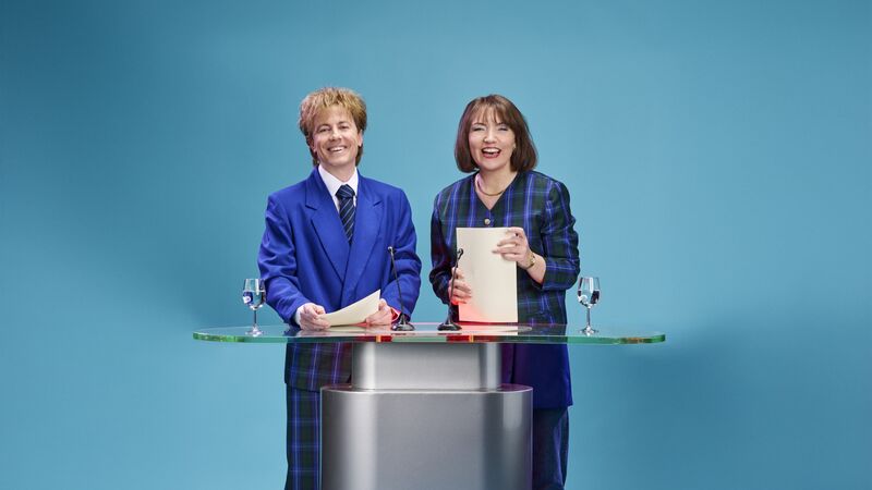 Twee vrouwen in formele kleding staan achter een glazen tafel op een blauwe achtergrond, lachend en met documenten in hun handen.