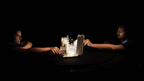 Twee mensen aan een tafel reiken naar een verlicht schaalmodel in een donkere omgeving.
