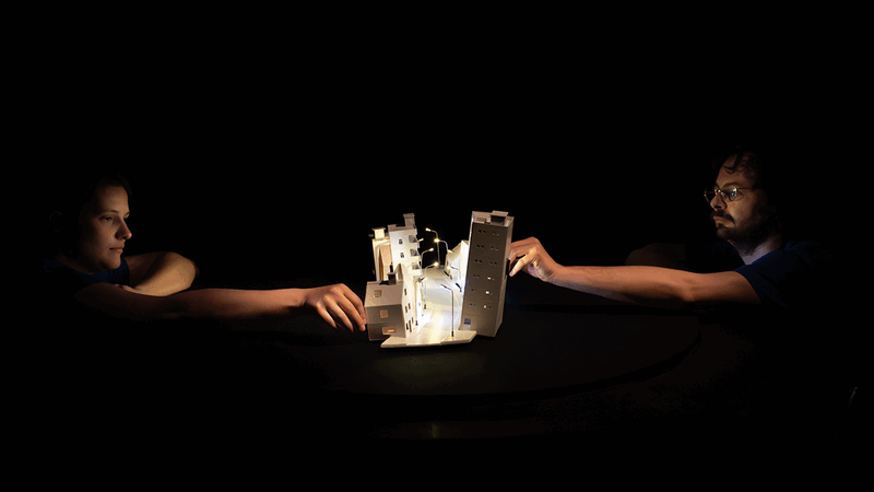 Twee mensen aan een tafel reiken naar een verlicht schaalmodel in een donkere omgeving.