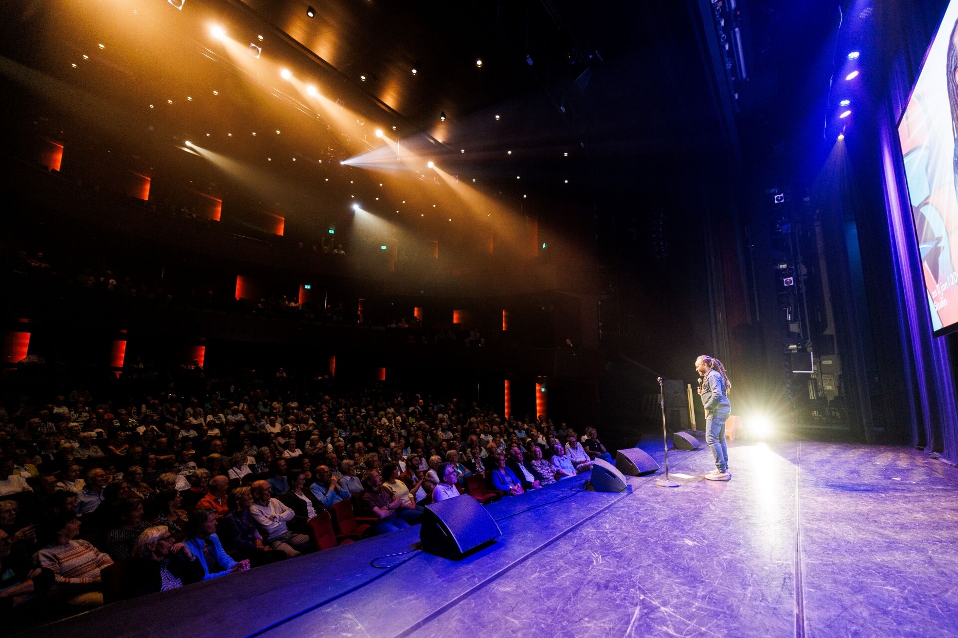 Performer op een podium voor een gevulde theaterzaal met fel gekleurde lichten