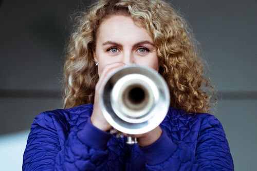 Vrouw met krullend haar houdt een trompet voor zich en kijkt recht in de camera.