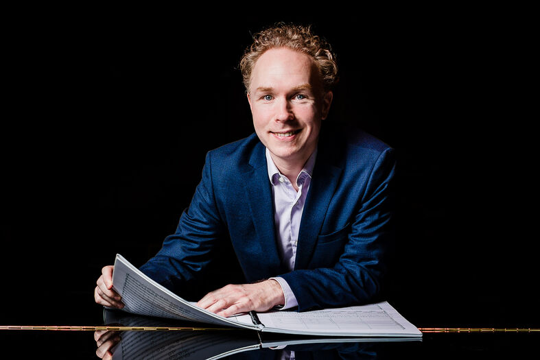 Man in een blauw pak zit aan een bureau met notities, kijkt recht in de camera. Zwarte achtergrond.