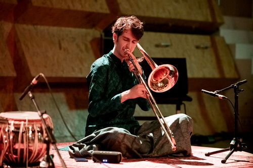 Muzikant speelt trombone zittend op de vloer, omgeven door muziekinstrumenten en microfoons.