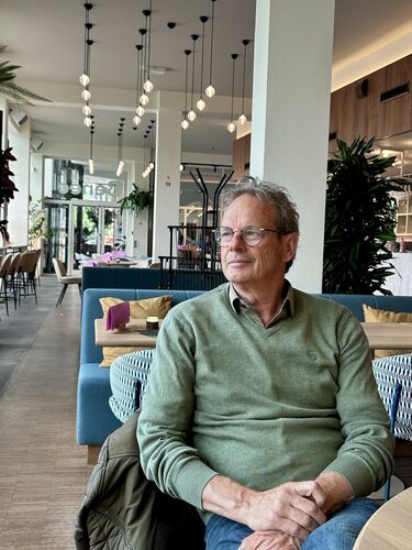 Een man met grijs haar in een groen sweater zit ontspannen in een modern café. Grote ramen en groen planten zijn zichtbaar.
