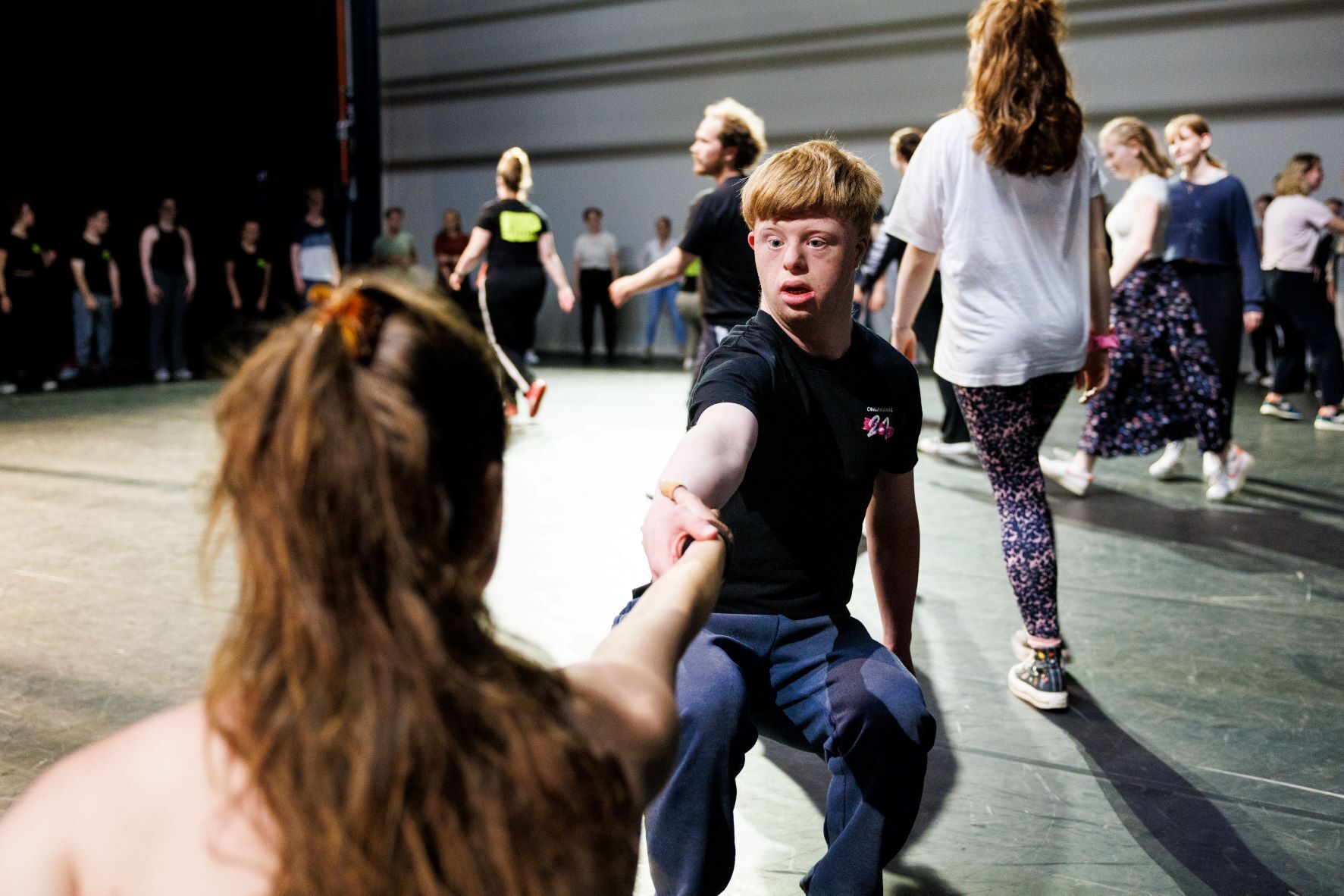 Groep dansers in een repetitie, met een jongen die een hand uitsteekt naar een meisje terwijl anderen achter hen aan het dansen zijn.