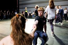 Groep dansers in een repetitie, met een jongen die een hand uitsteekt naar een meisje terwijl anderen achter hen aan het dansen zijn.