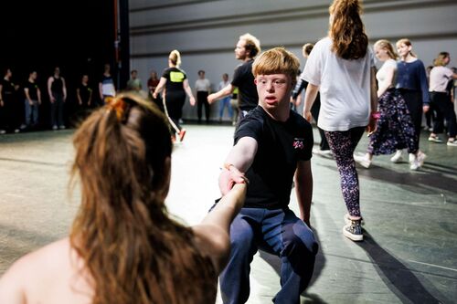 Groep dansers in een repetitie, met een jongen die een hand uitsteekt naar een meisje terwijl anderen achter hen aan het dansen zijn.