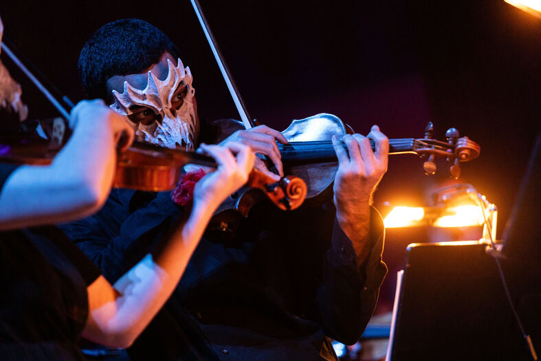 Stringplayers in zwarte kleding, met maskers, spelen violen in een sfeervolle omgeving met warme verlichting.