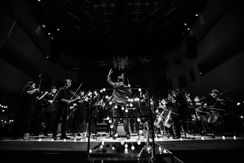 Zwart-wit afbeelding van een orkest op het podium, met een dirigent die de muzikanten aanstuurt.