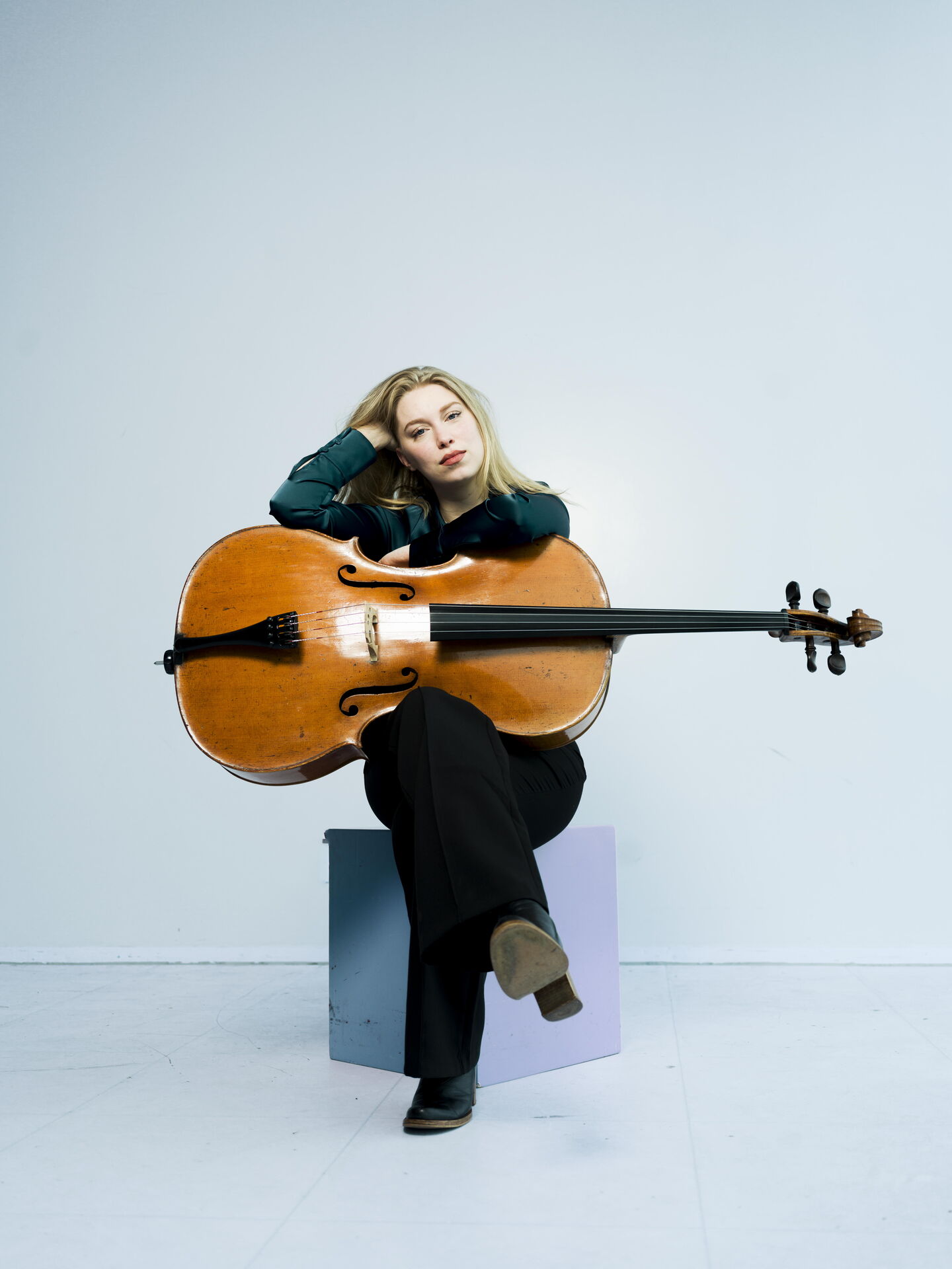 Vrouw met een cello zittend op een stoel, leunt achterover met een hand in haar haar, tegen een lichte achtergrond.
