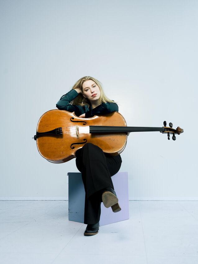 Vrouw met een cello zittend op een stoel, leunt achterover met een hand in haar haar, tegen een lichte achtergrond.