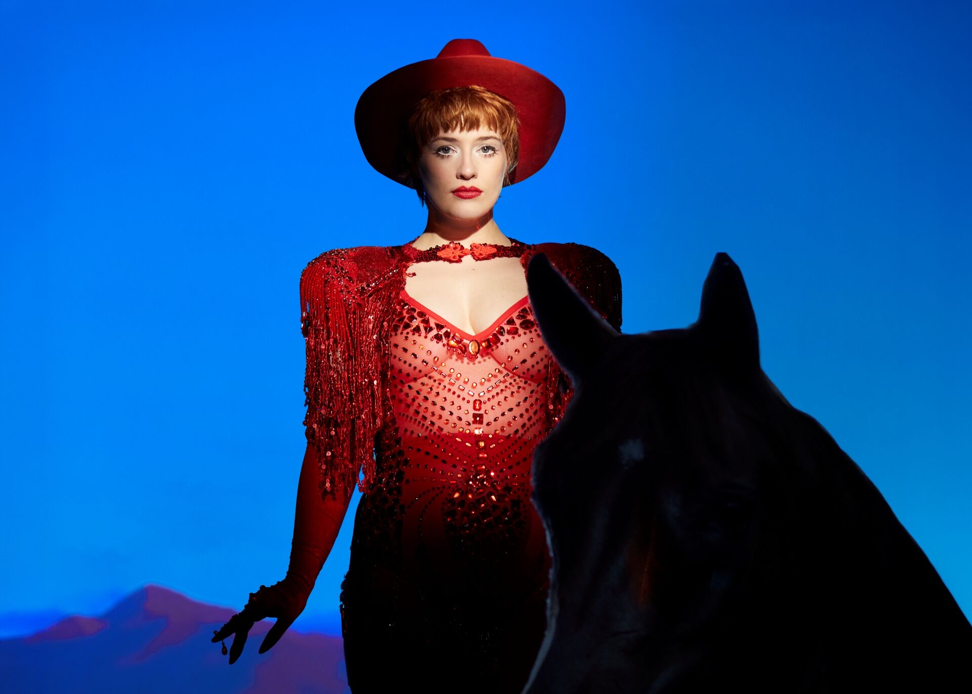 Vrouw in glinsterende rode western‑outfit en hoed staat voor een blauwe achtergrond, met de donkere silhouetvorm van een paard op de voorgrond.