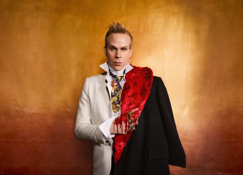 Een man in een elegantere outfit met een zwart jasje en een rode sjaal, staand voor een gouden achtergrond.