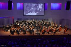Een orkest speelt op het podium, met een groot scherm op de achtergrond dat historische beelden toont.