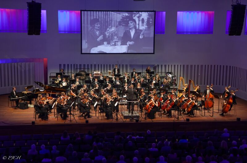 Een orkest speelt op het podium, met een groot scherm op de achtergrond dat historische beelden toont.